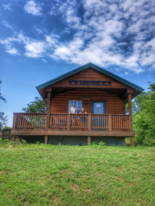 A wooden cabin with a porch, surrounded by greenery and a blue sky, featuring a person standing at the entrance.